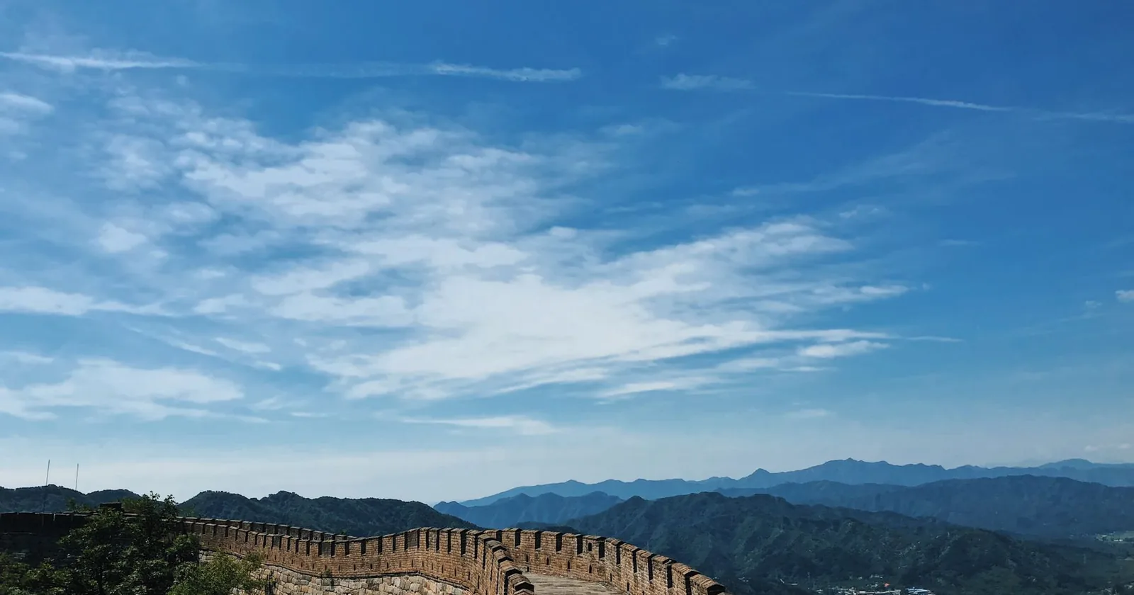 A restored, crenellated section of the Great Wall of China curving along a forested ridge, with layered blue mountain ranges receding under a wide sky.