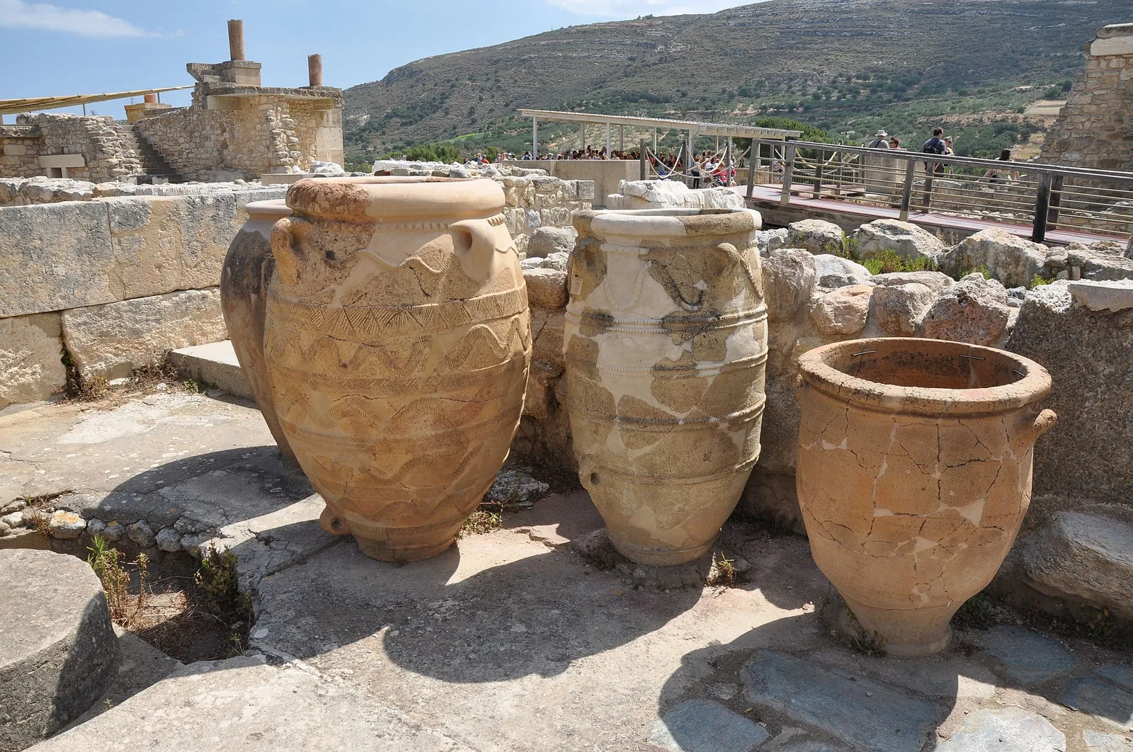 Three giant decorated pithoi, Minoan storage jars, standing among the ruins of Knossos with visitors on a walkway behind.