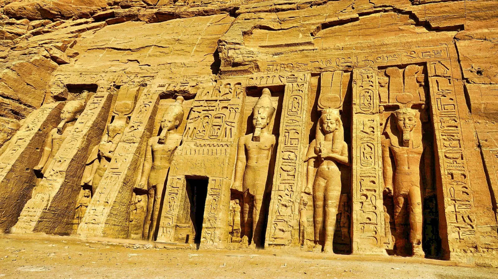 The rock-cut facade of the Small Temple at Abu Simbel, with six standing colossi of Ramesses II and Nefertari set in niches between buttresses covered in hieroglyphic inscriptions.