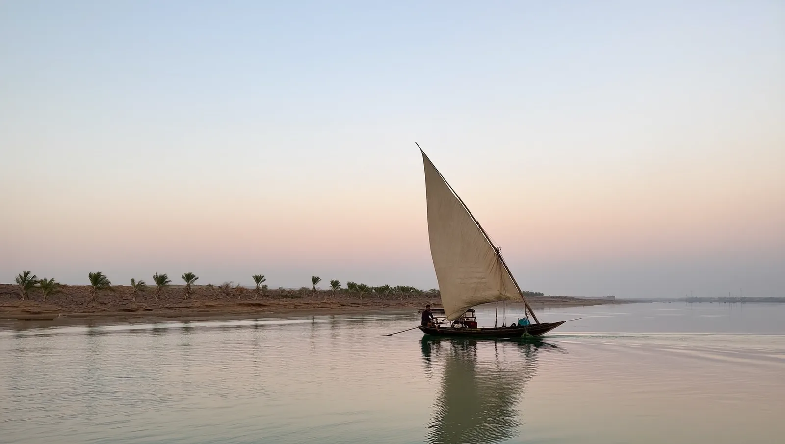 A traditional wooden boat with a single tall triangular sail on a calm river at dawn, with palms along the far bank and pink light on glassy water.