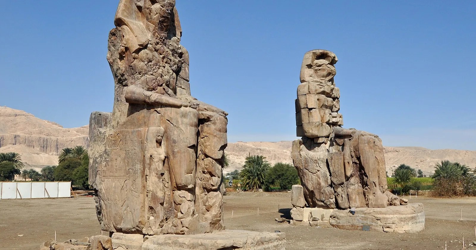 The two seated Colossi of Memnon, weathered statues of Amenhotep III, rising from the plain of the Luxor west bank below distant desert cliffs.