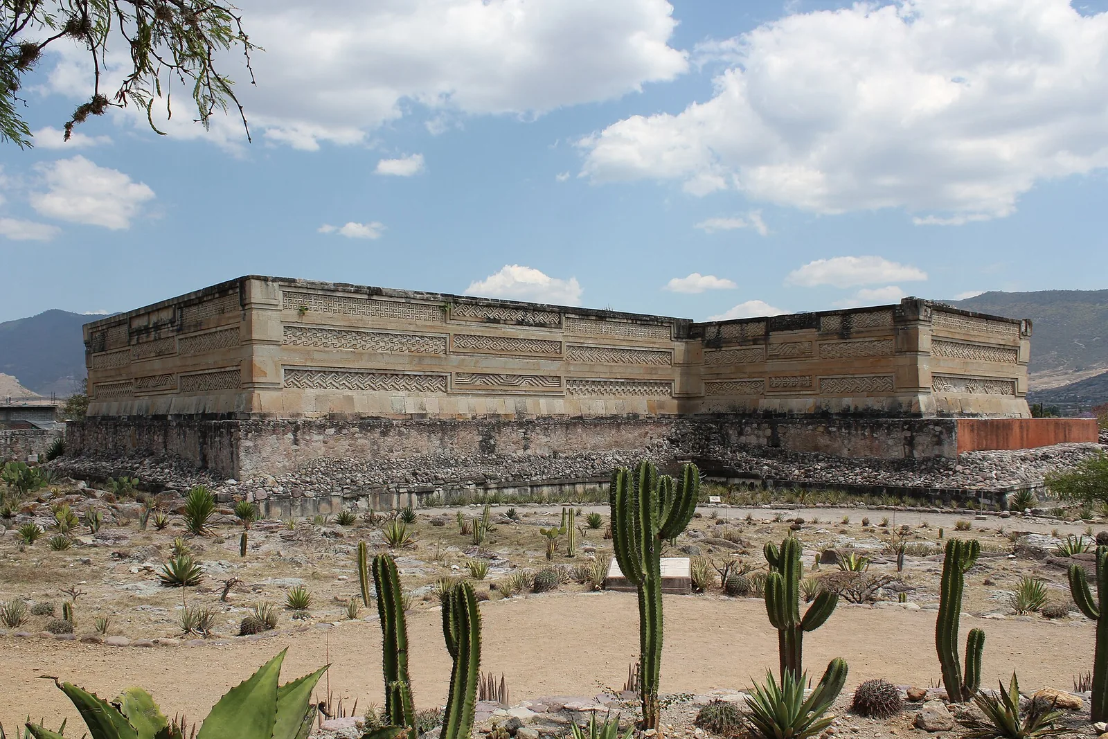 The long stone facade of the Palace of Columns at Mitla, its upper walls covered in bands of geometric fretwork, with cactus and agave in the foreground.