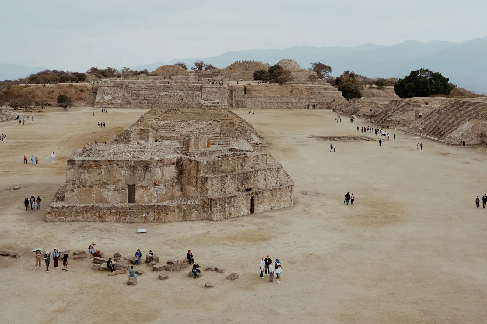 A wide view across Monte Alban's Great Plaza, with visitors crossing the flat open ground between low stone platforms and mountains on the horizon.