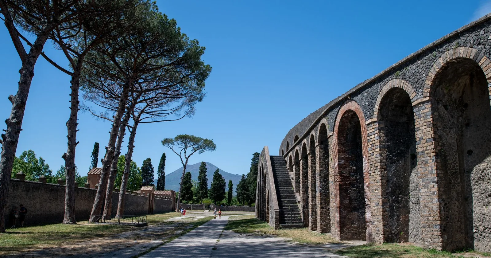 The arched stone exterior of Pompeii's amphitheatre beside umbrella pines, with Mount Vesuvius rising in the distance.