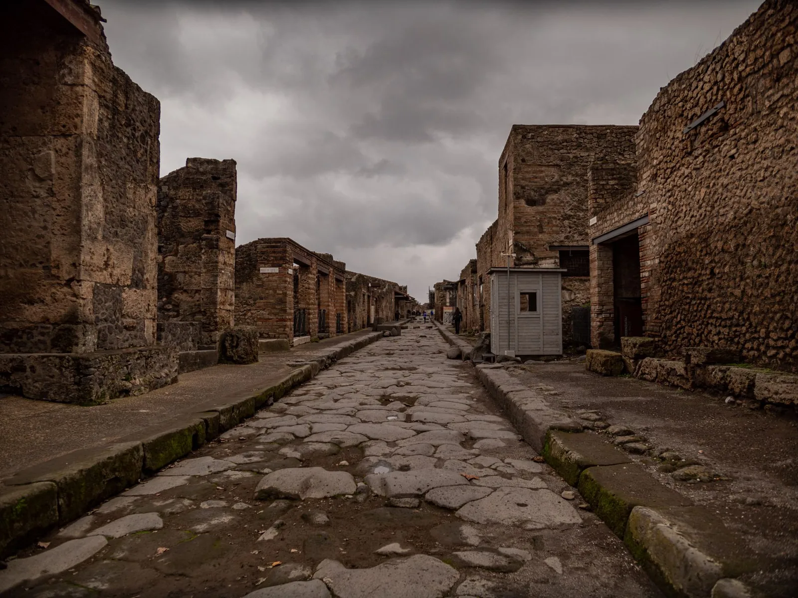 A deserted ancient street in Pompeii lined with weathered stone ruins under an overcast sky.