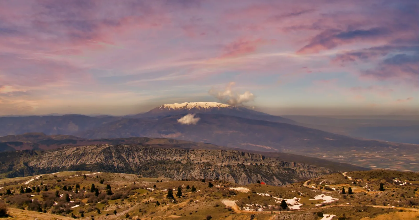 The snow-capped summit ridge of Mount Olympus rising above the Greek coastal plain under a pink sunset sky.