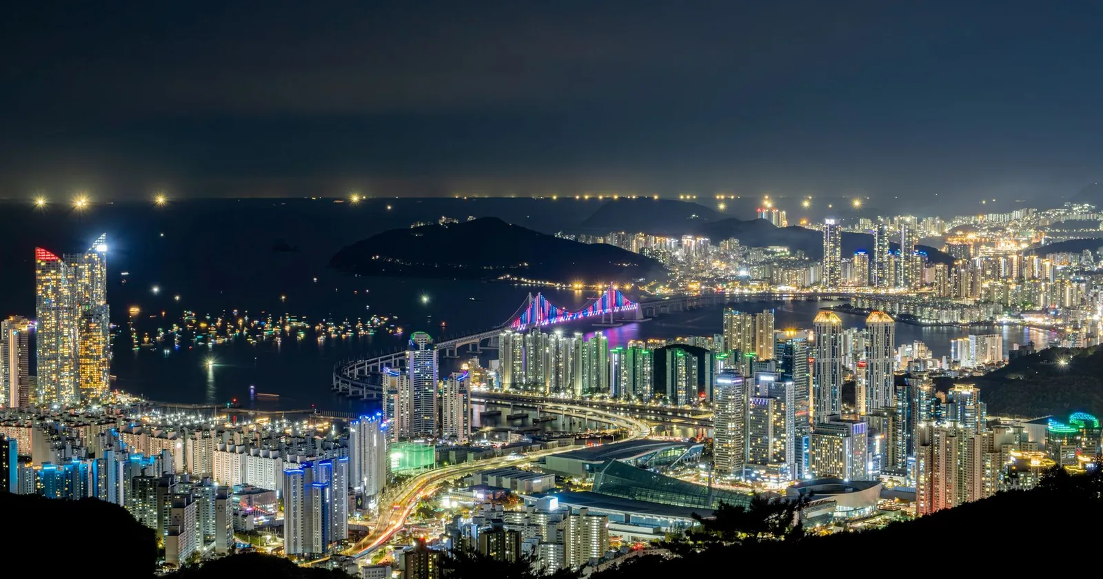 Busan's illuminated night skyline with the lit Gwangan Bridge curving across the bay toward high-rise districts.