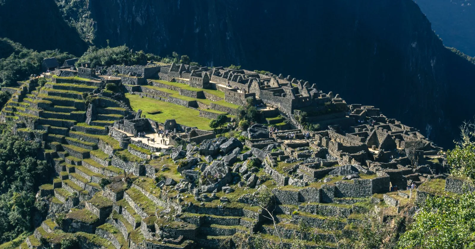 Aerial view of the Inca citadel of Machu Picchu, its stone terraces and roofless buildings spread along a high ridge above a dark valley.