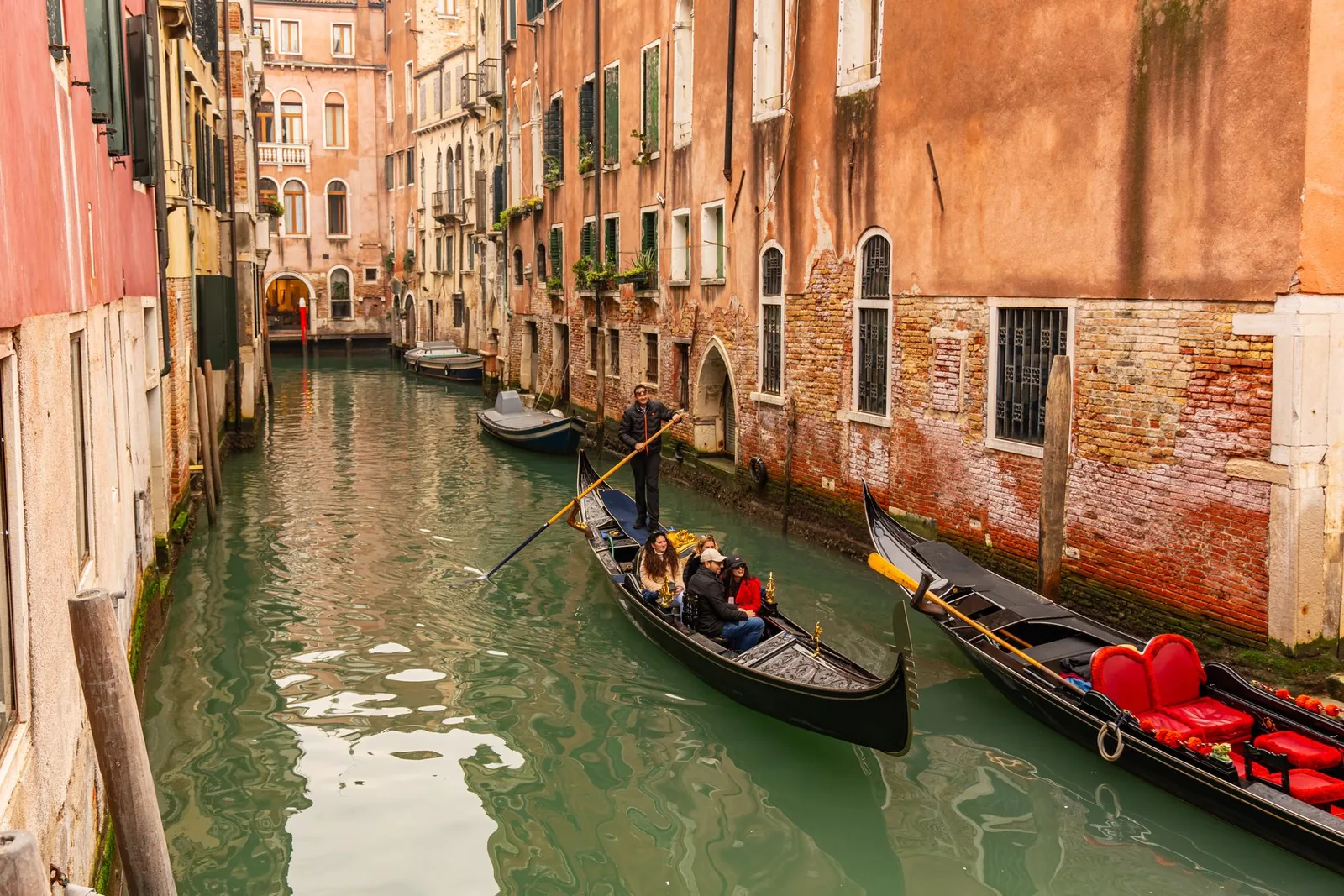 Two gondolas carrying passengers along a narrow Venetian canal between weathered brick and plaster buildings.